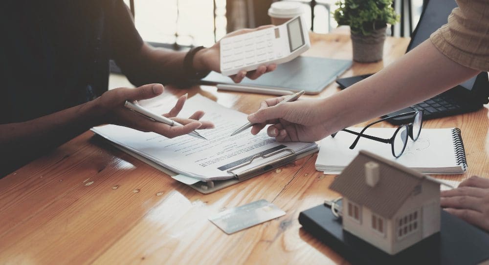 Real estate agent assisting client to sign contract paper at desk with house model