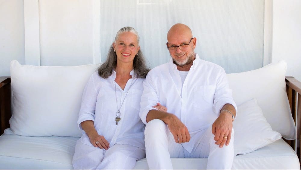 A friendly portrait of Bettina and Ralph from Team Haverkate, an older couple with gray hair, dressed in white, sitting closely on a white couch and smiling at the camera.