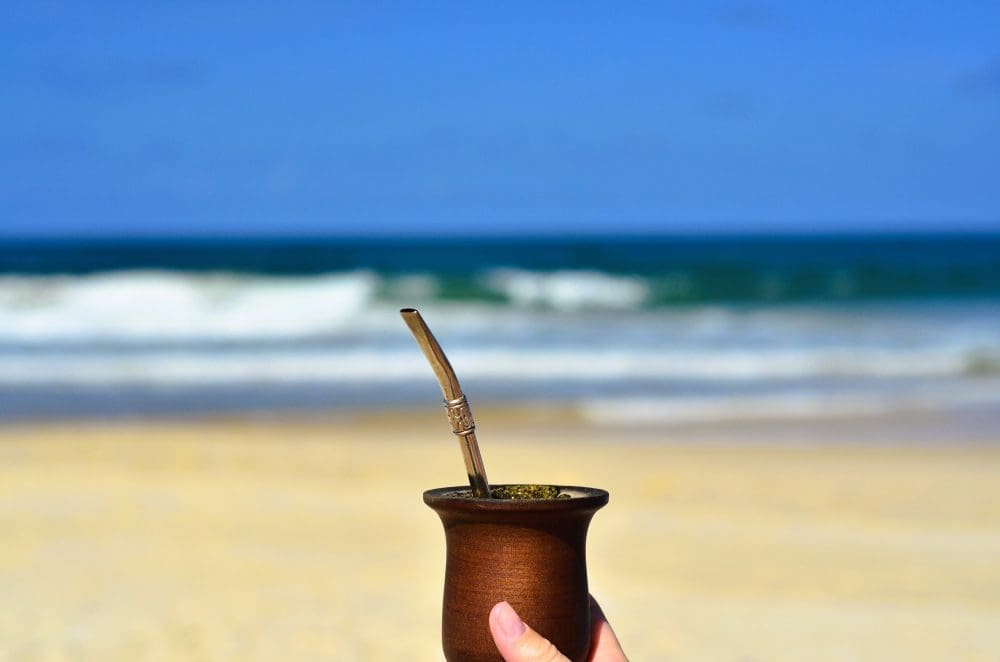 Eine Hand hält eine traditionelle Mate-Kalebasse mit einer Bombilla an einem Sandstrand, mit Meereswellen im Hintergrund.