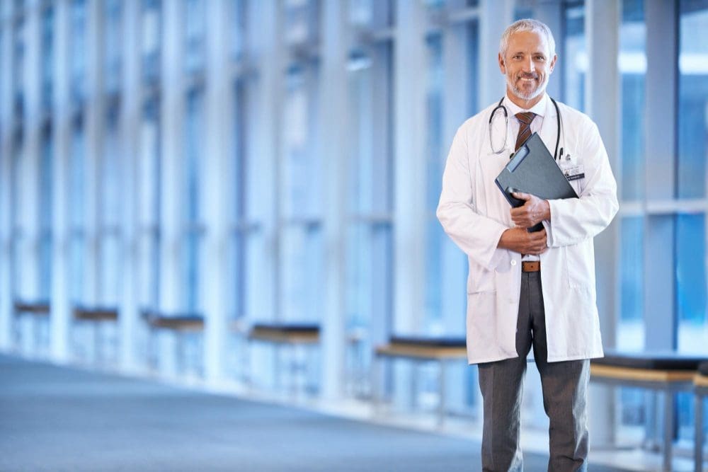 A smiling male doctor, with gray hair and a beard, wearing a white lab coat and stethoscope, stands confidently in a bright, modern hospital hallway, holding a clipboard.
