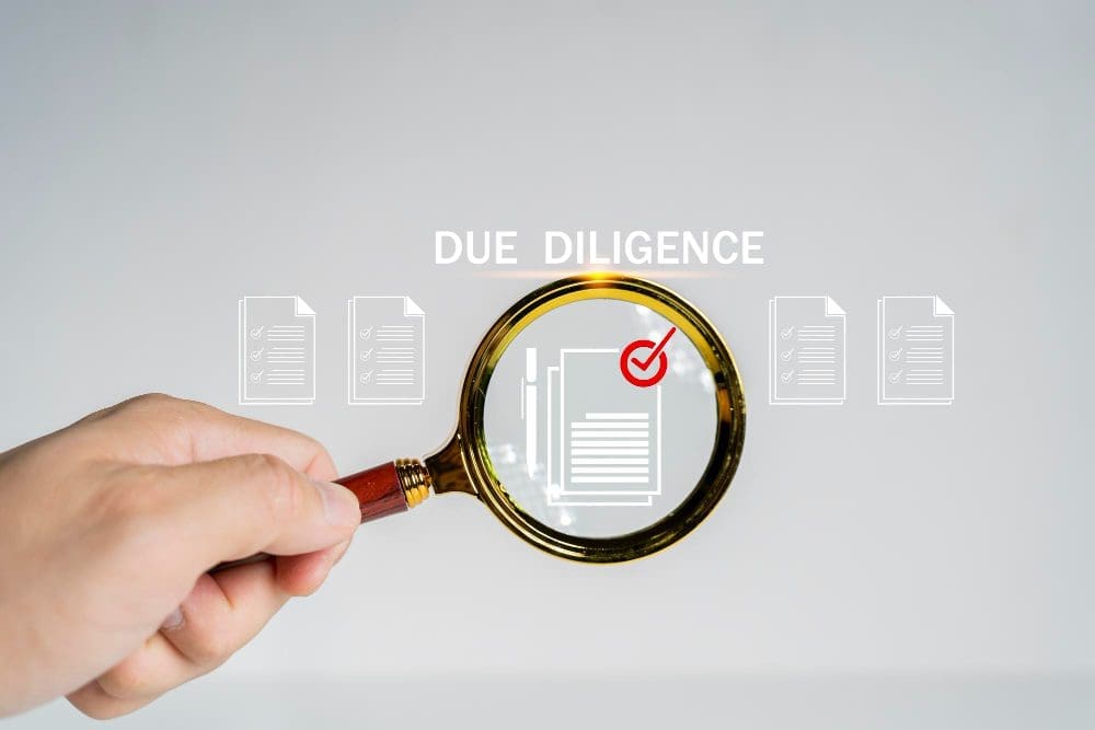 A hand holds a magnifying glass over a document icon with a red checkmark, surrounded by other document icons, and the text "DUE DILIGENCE" above, against a plain grey background.