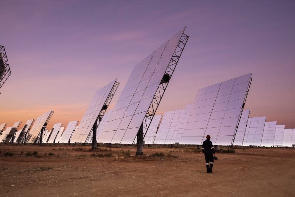 A vast solar power plant at sunset or sunrise, with rows of large, reflective solar panels angled towards the sky. A lone figure of a worker walks in the foreground, highlighting the scale of the renewable energy installation.