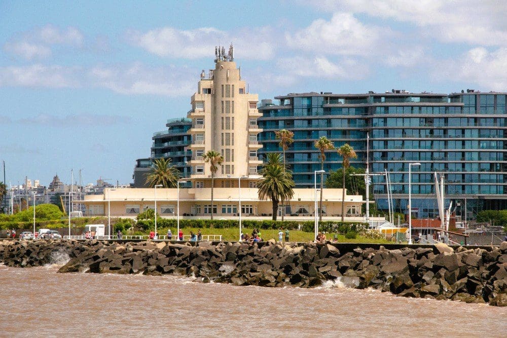 Ein Blick auf die Uferpromenade von Montevideo mit dem markanten beigen Gebäude des uruguayischen Yachtclubs mit einem Turm im Vordergrund und dem modernen blauen Forum-Gebäude im Hintergrund, alles hinter einem felsigen Wellenbrecher.