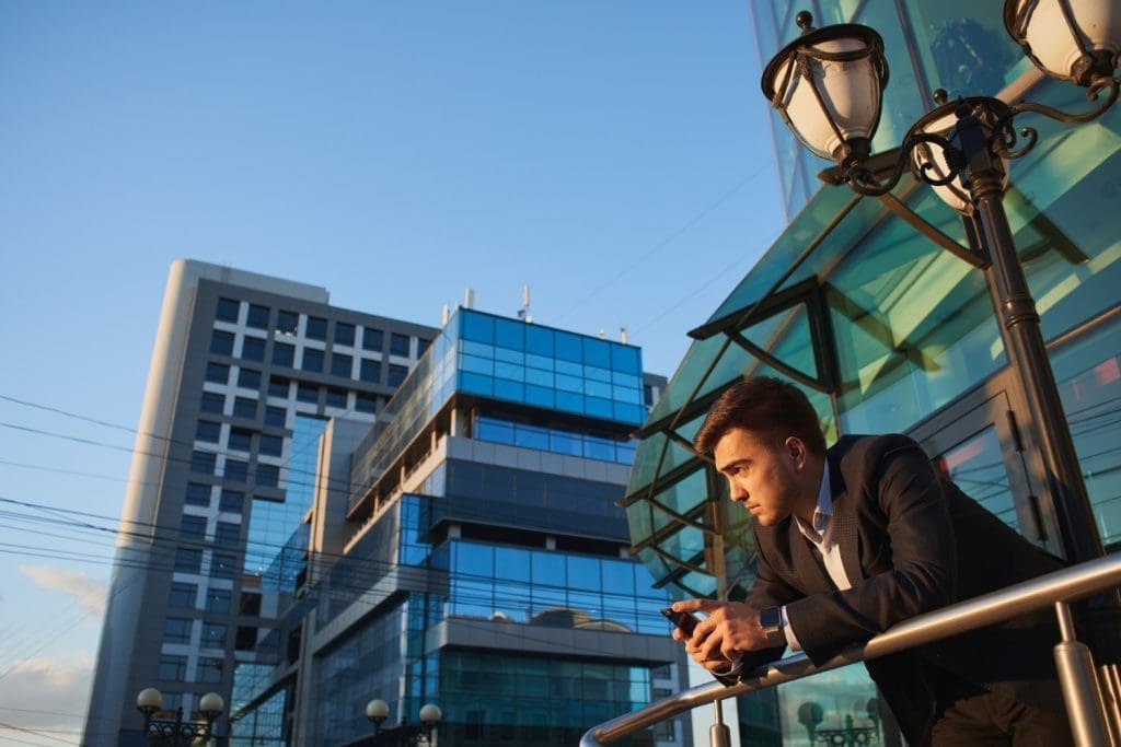 A man in a dark suit leans on a metal railing and looks at his smartphone, with modern office buildings and a bright blue sky in the background.