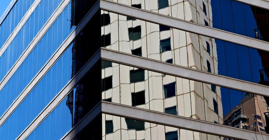 Corner of a modern building facade with blue reflective glass showing mirrored reflections of nearby structures.