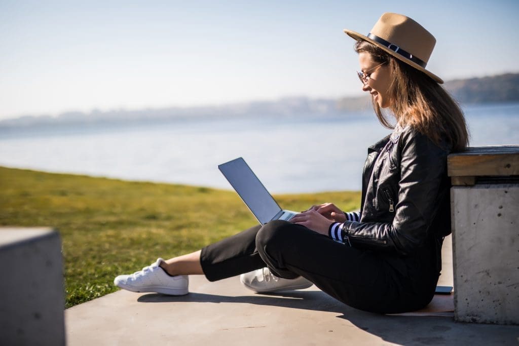 Junge Frau mit Hut und Lederjacke arbeitet im Freien an einem Laptop in der Nähe eines Gewässers.