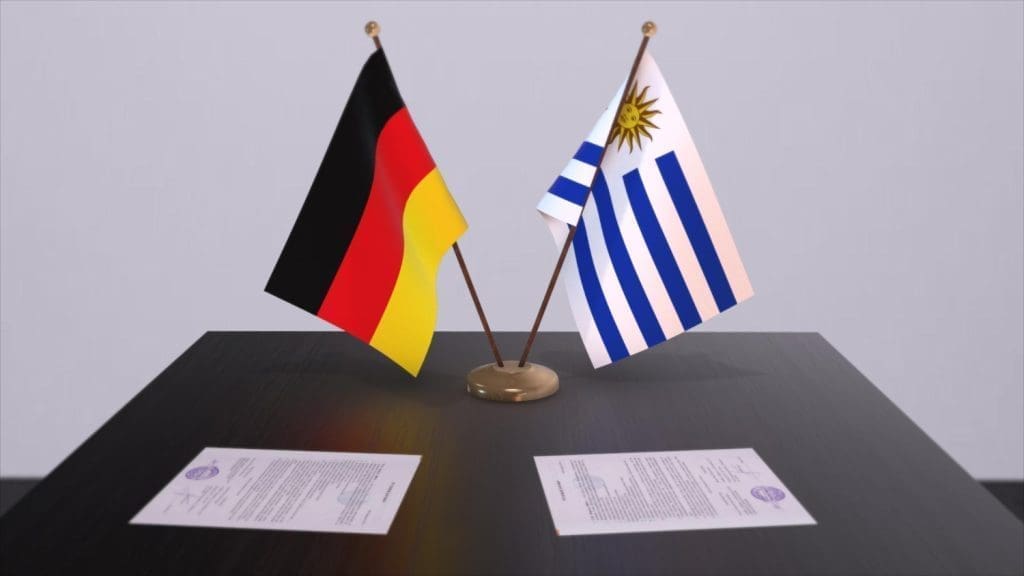 The flags of Germany and Uruguay stand side-by-side on a desk with two contracts lying underneath them.