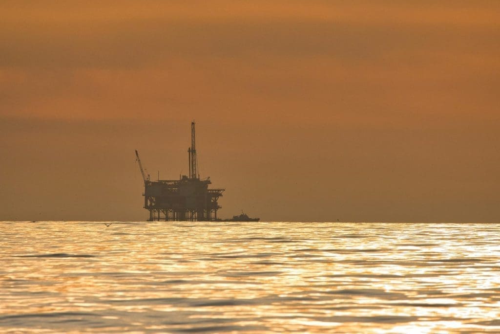 Silhouette einer Offshore-Ölbohrinsel mit einem kleinen Boot in der Nähe auf einem schimmernden goldenen Meer unter einem orangefarbenen Himmel bei Sonnenuntergang oder Sonnenaufgang.