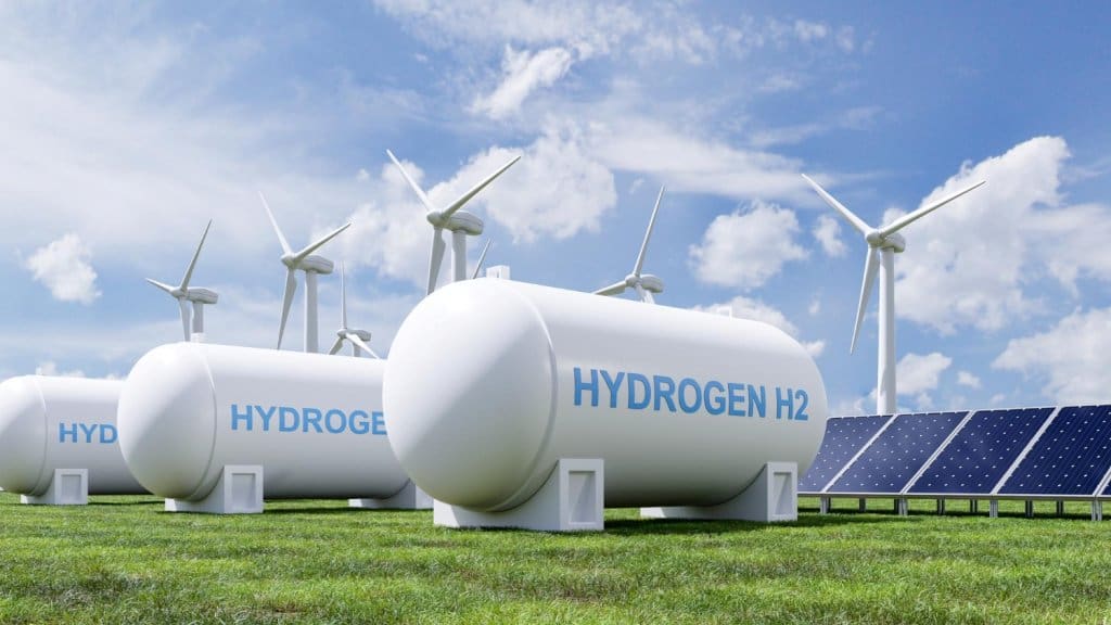 White storage tanks labeled Hydrogen H2 are powered by nearby wind turbines and solar arrays in a bright grassy field.