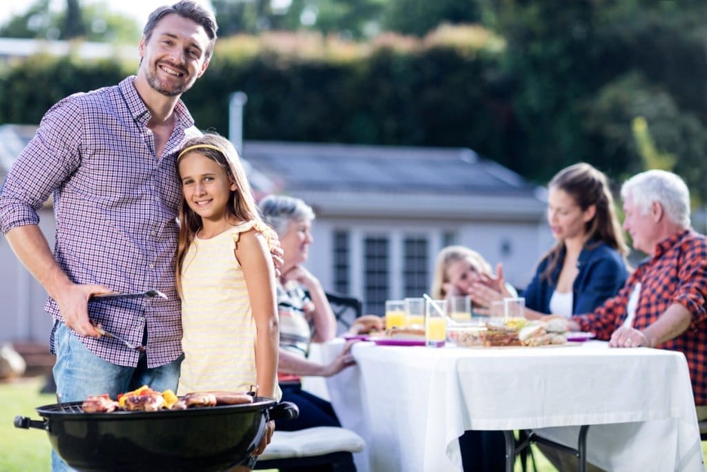 Smiling father and daughter stand by a barbecue while family dines outdoors in the sunny backyard.