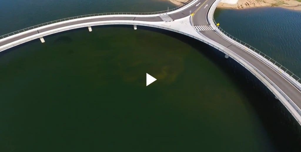 Luftaufnahme einer modernen, geschwungenen Brückenkonstruktion, die dunkelgrünes Wasser nahe einer felsigen Küste überspannt.